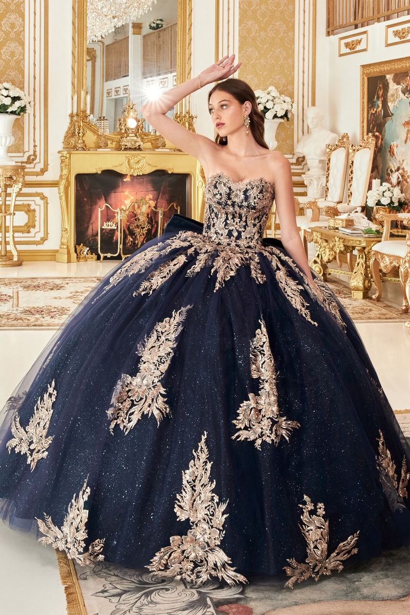 A woman models the Ladivine Strapless Quinceanera Dress 15715—a luxurious gold and navy ball gown—in an ornate, gold-accented room with a fireplace and elegant decor.